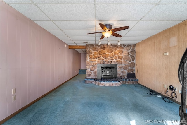 unfurnished living room featuring a drop ceiling, carpet floors, wood walls, a ceiling fan, and a wood stove