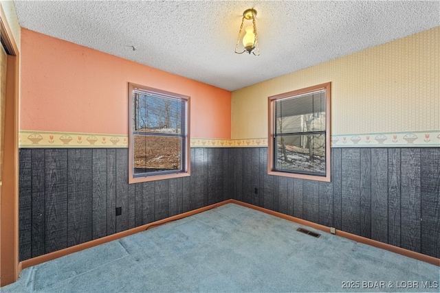 carpeted empty room with a textured ceiling, wainscoting, visible vents, and wallpapered walls