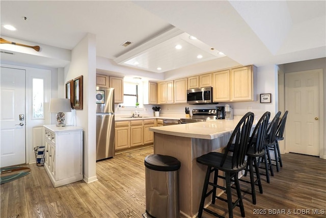 kitchen with stainless steel appliances, light brown cabinets, light wood-type flooring, a peninsula, and a kitchen bar