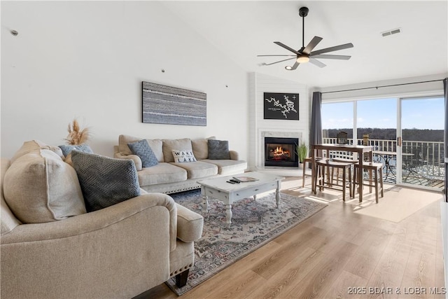 living area featuring visible vents, a glass covered fireplace, ceiling fan, wood finished floors, and vaulted ceiling