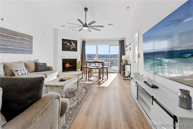 living room with lofted ceiling, visible vents, light wood-style flooring, a ceiling fan, and a warm lit fireplace