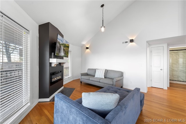 living room featuring high vaulted ceiling, a baseboard radiator, light wood-style flooring, visible vents, and a glass covered fireplace