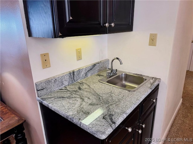 kitchen featuring carpet flooring, a sink, light stone countertops, dark cabinetry, and baseboards