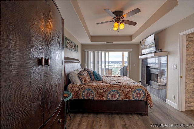 bedroom with baseboards, a tiled fireplace, ceiling fan, wood finished floors, and a tray ceiling