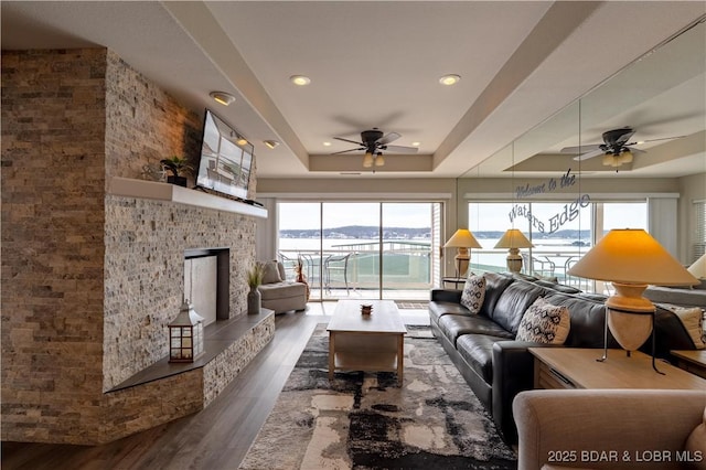 living area with ceiling fan, a tray ceiling, a fireplace with raised hearth, and dark wood-style flooring