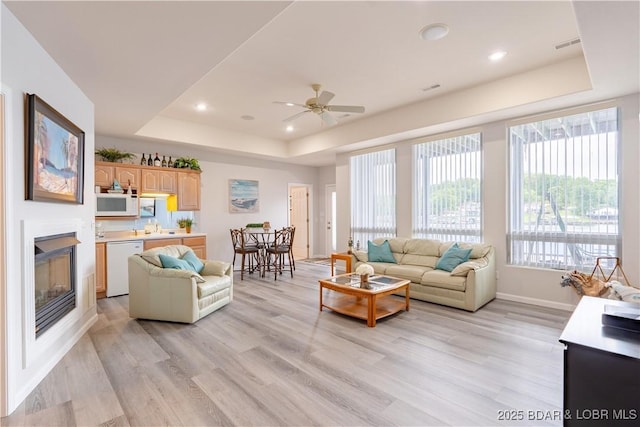 living area with a glass covered fireplace, light wood-type flooring, a raised ceiling, and baseboards