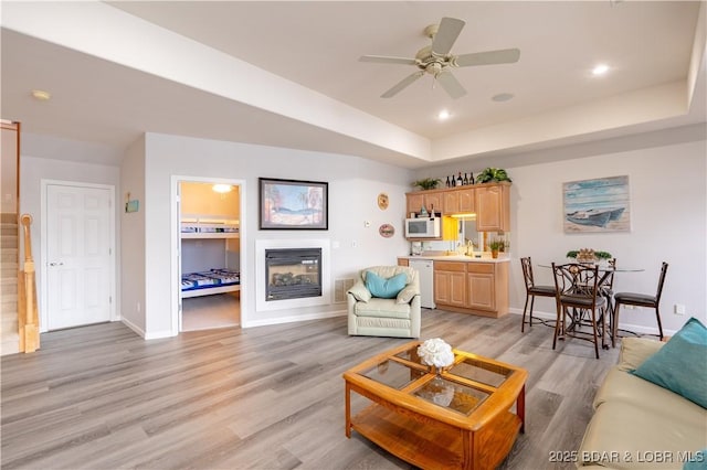 living area featuring recessed lighting, baseboards, light wood-style floors, a raised ceiling, and a glass covered fireplace