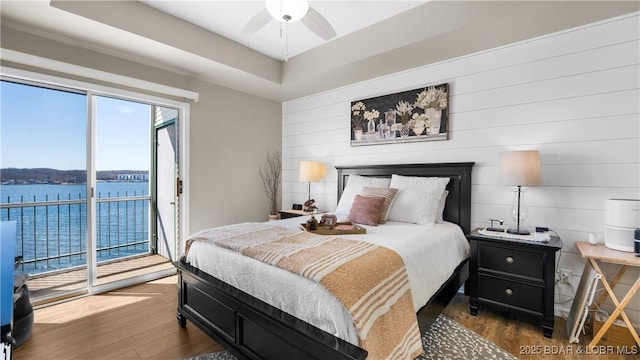 bedroom with a raised ceiling, ceiling fan, wood finished floors, a water view, and access to exterior