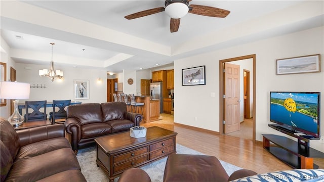 living room featuring light wood-type flooring, a raised ceiling, baseboards, and ceiling fan with notable chandelier