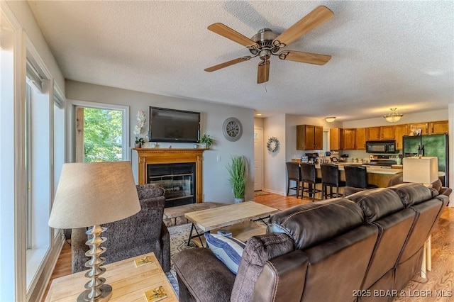 living area with a textured ceiling, light wood finished floors, a glass covered fireplace, and a ceiling fan