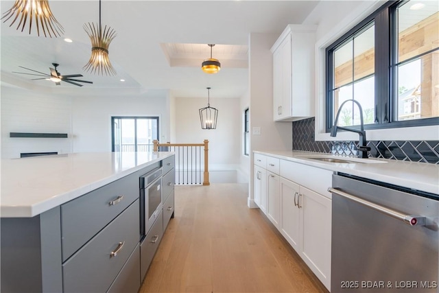 kitchen with gray cabinetry, a sink, backsplash, stainless steel dishwasher, and a raised ceiling