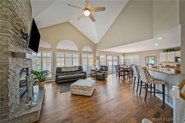 living room featuring ceiling fan with notable chandelier, high vaulted ceiling, dark wood-style flooring, and a fireplace