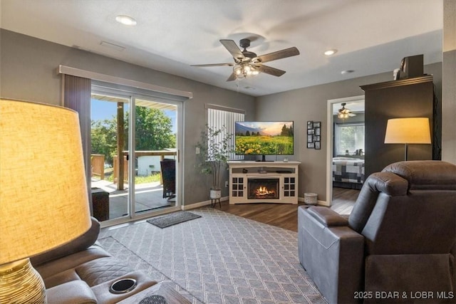 living room featuring a warm lit fireplace, plenty of natural light, a ceiling fan, and wood finished floors