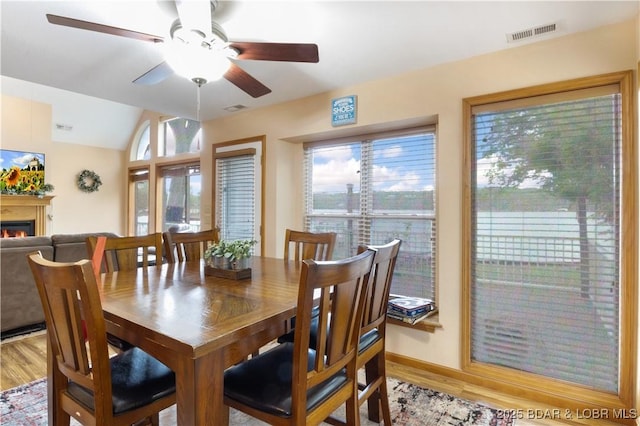 dining space featuring ceiling fan, visible vents, vaulted ceiling, light wood finished floors, and a glass covered fireplace