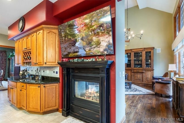 kitchen with a chandelier, high vaulted ceiling, a multi sided fireplace, dark stone counters, and light wood finished floors