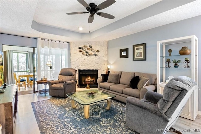 living room with a fireplace, light wood finished floors, a raised ceiling, a ceiling fan, and a textured ceiling