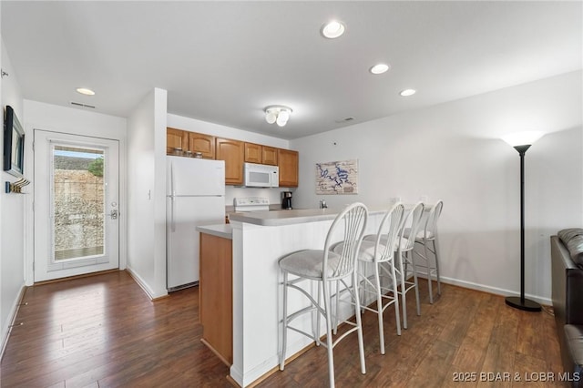 kitchen with a kitchen bar, brown cabinets, a peninsula, dark wood-style floors, and white appliances