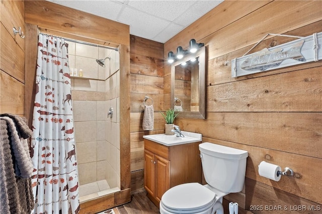 bathroom with a stall shower, wooden walls, a drop ceiling, toilet, and vanity