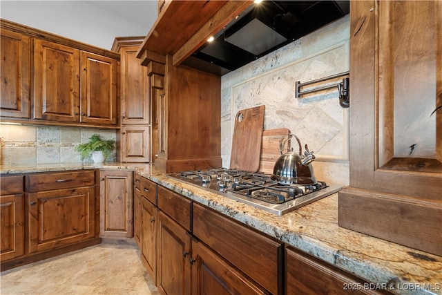 kitchen featuring premium range hood, light stone counters, backsplash, stainless steel gas stovetop, and brown cabinetry