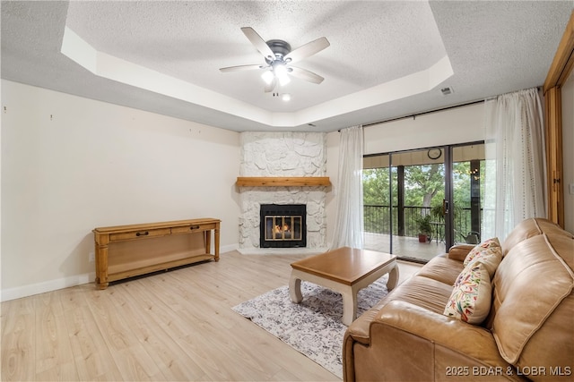 living room with a textured ceiling, a stone fireplace, a tray ceiling, and wood finished floors