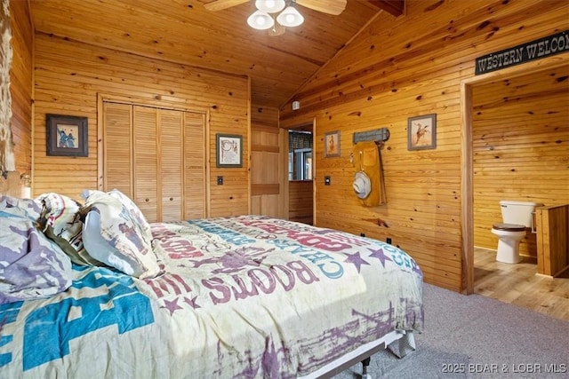 carpeted bedroom with vaulted ceiling with beams, ceiling fan, a closet, ensuite bathroom, and wood walls