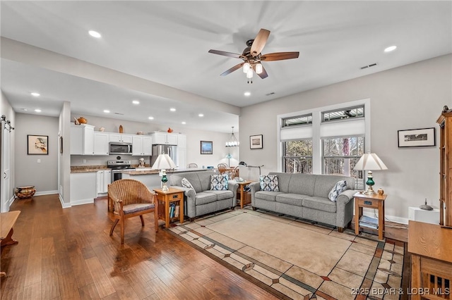 living area featuring a barn door, ceiling fan with notable chandelier, dark wood-style flooring, and baseboards