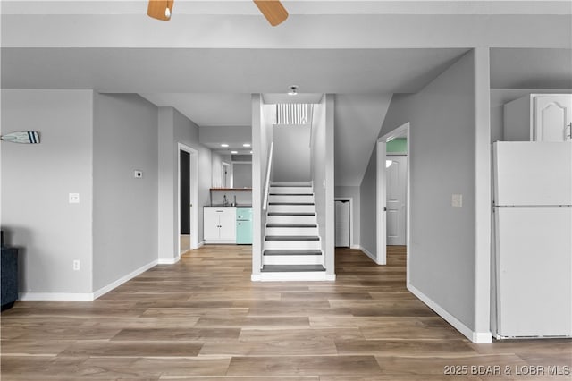 unfurnished living room featuring baseboards, stairs, a ceiling fan, and light wood finished floors