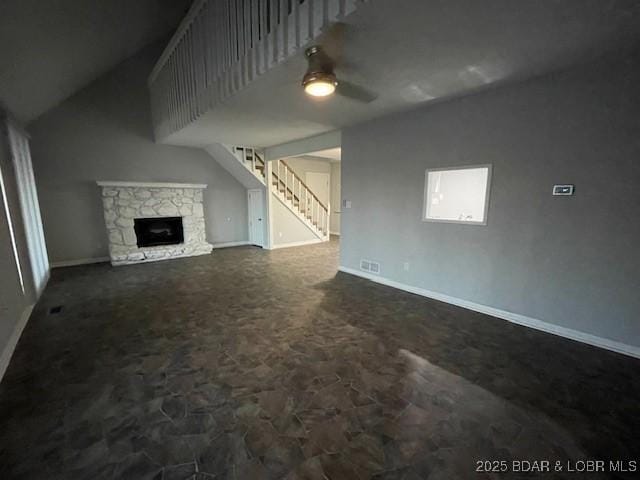 unfurnished living room with baseboards, stairway, a stone fireplace, a ceiling fan, and dark colored carpet