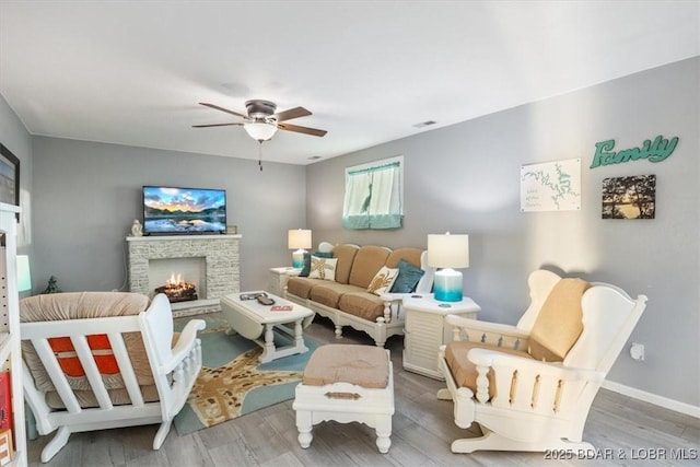 living room featuring ceiling fan, baseboards, a warm lit fireplace, and wood finished floors