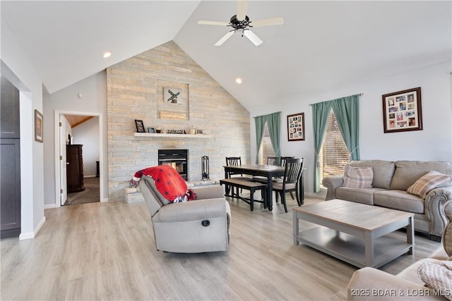 living room with high vaulted ceiling, light wood-style flooring, a ceiling fan, a stone fireplace, and baseboards