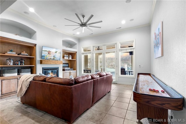 living room with built in features, light tile patterned flooring, ceiling fan, ornamental molding, and a glass covered fireplace