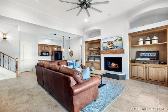 living room featuring crown molding, ceiling fan, built in features, stairway, and a warm lit fireplace