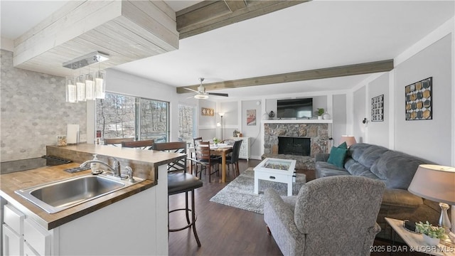 living room with dark wood-style floors, beam ceiling, a stone fireplace, and ceiling fan