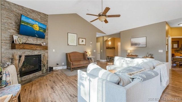 living room featuring wood finished floors, baseboards, a ceiling fan, arched walkways, and a stone fireplace