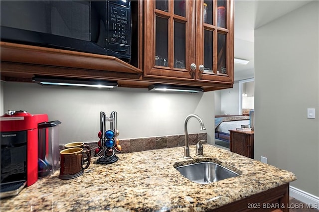 kitchen with a sink, light stone counters, black microwave, glass insert cabinets, and baseboards