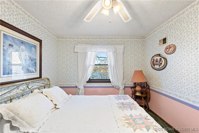 bedroom with visible vents, a textured ceiling, wainscoting, wallpapered walls, and ceiling fan