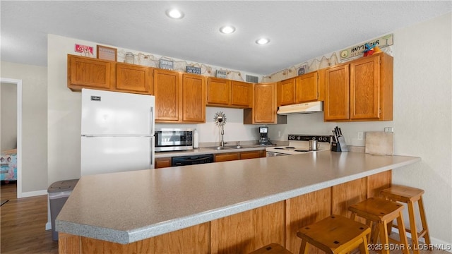 kitchen featuring white appliances, a peninsula, a sink, under cabinet range hood, and brown cabinets