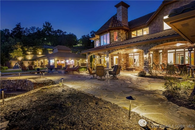 back of house at night featuring a gazebo, a chimney, a patio area, and stone siding