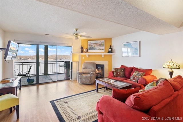 living area featuring visible vents, wood finished floors, a glass covered fireplace, a textured ceiling, and a ceiling fan