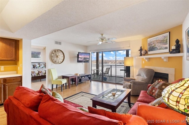 living area featuring a ceiling fan, visible vents, light wood finished floors, a fireplace, and a textured ceiling