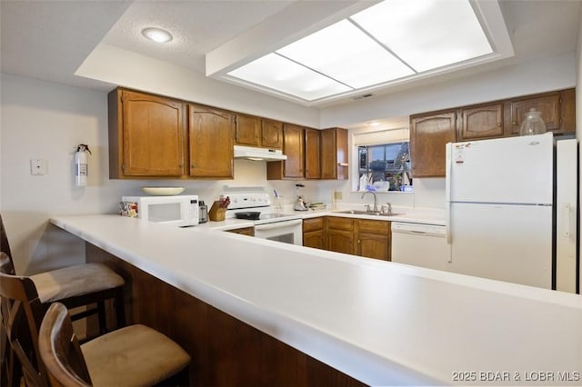 kitchen with under cabinet range hood, light countertops, brown cabinetry, white appliances, and a sink