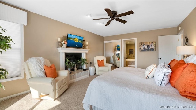 bedroom featuring a ceiling fan, baseboards, ensuite bath, light carpet, and a glass covered fireplace