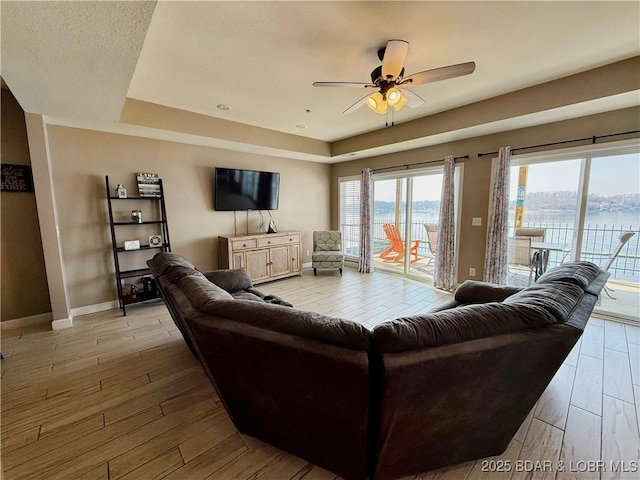 living area featuring a raised ceiling, a ceiling fan, a textured ceiling, light wood finished floors, and baseboards