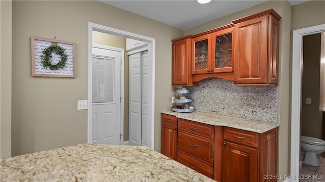 kitchen with light stone counters, decorative backsplash, brown cabinetry, and glass insert cabinets