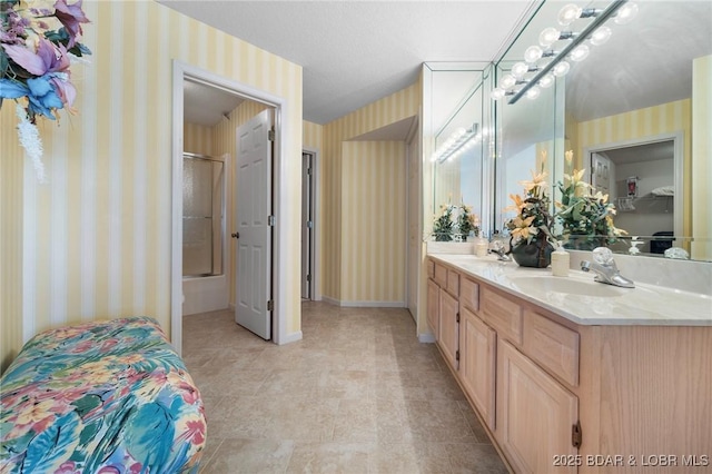 full bath featuring wallpapered walls, baseboards, double vanity, combined bath / shower with glass door, and a sink