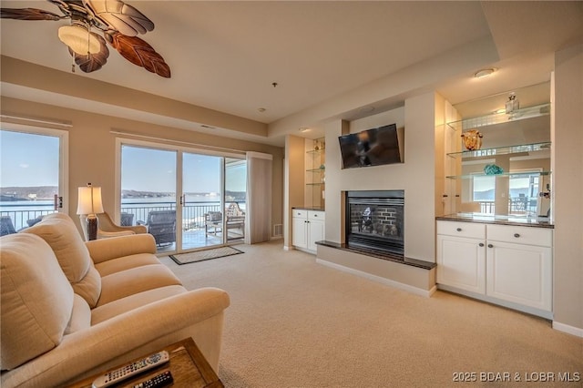 living room featuring built in shelves, baseboards, ceiling fan, light carpet, and a glass covered fireplace