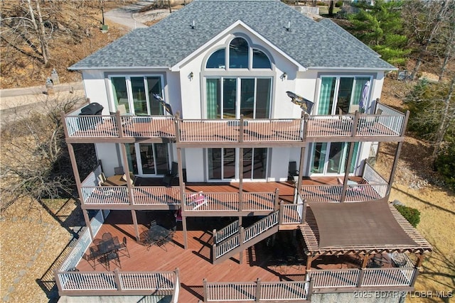 rear view of property featuring stucco siding, a balcony, a wooden deck, and a shingled roof