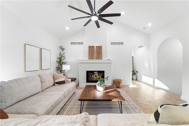 living room with arched walkways, a ceiling fan, wood finished floors, a large fireplace, and high vaulted ceiling