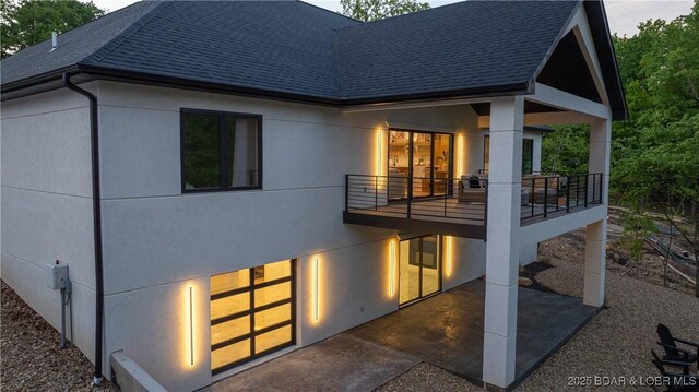 back of house featuring a shingled roof, a balcony, stucco siding, and a patio area