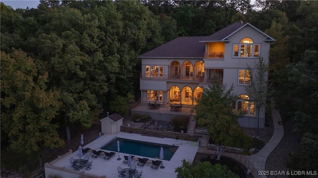 rear view of property with an outdoor structure, stucco siding, a balcony, a forest view, and a patio area